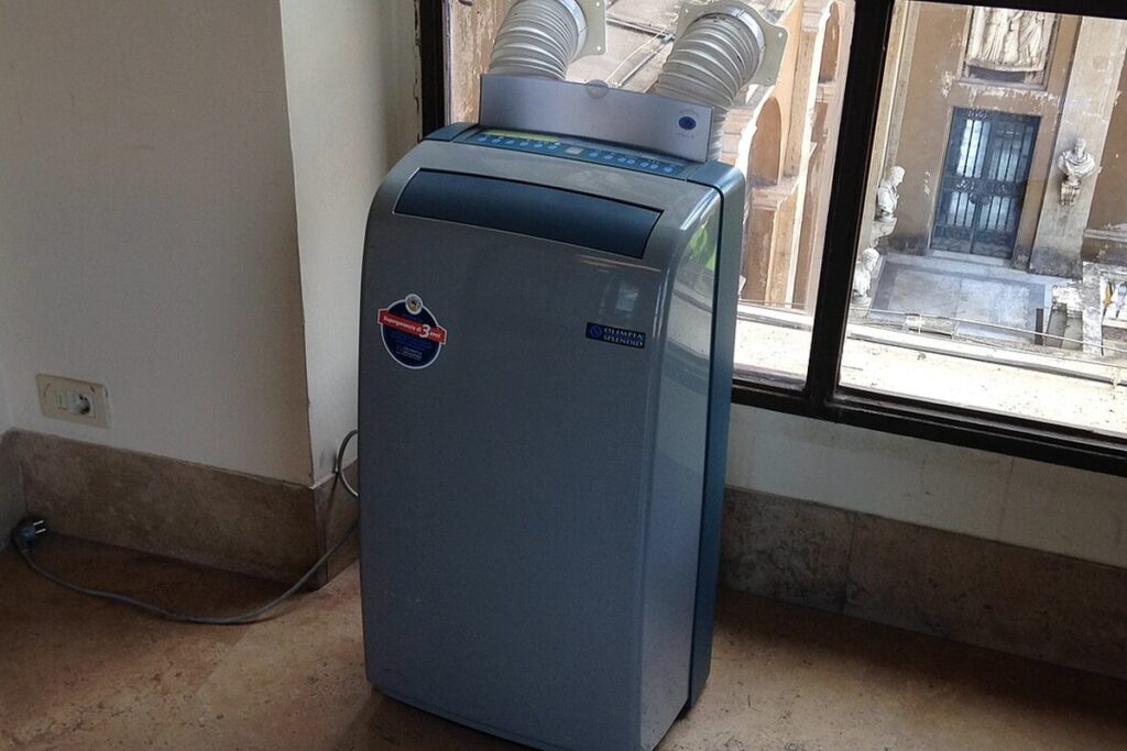 Climatiseur mobile 2026 : bien choisir (BTU réels, pièges) 2 Climatiseur mobile bleu près d une fenêtre, tuyaux d évacuation vers l extérieur